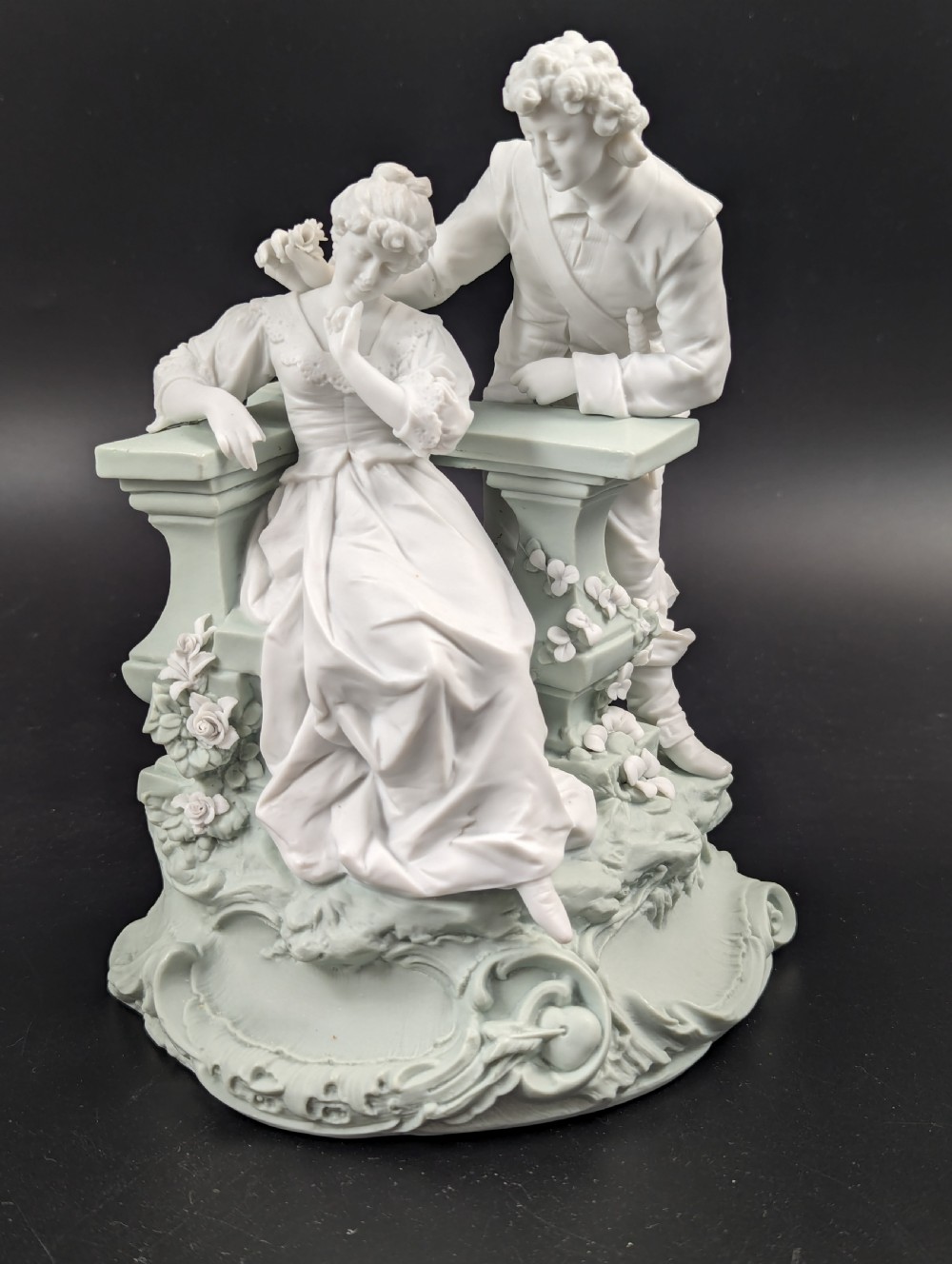 A white and pale green parian porcelain figurine depicting a courtly man and woman interacting beside a balustrade.