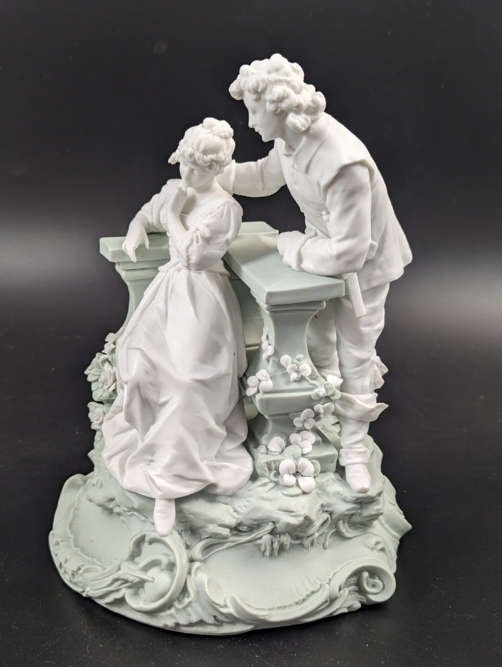 A white and pale green parian porcelain figurine depicting a courtly man and woman interacting beside a balustrade. View II