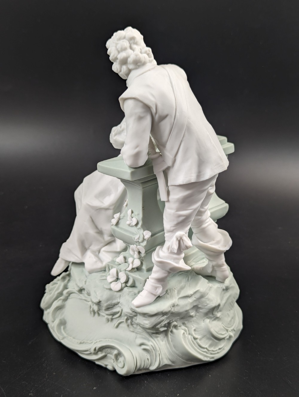 A white and pale green parian porcelain figurine depicting a courtly man and woman interacting beside a balustrade. View III