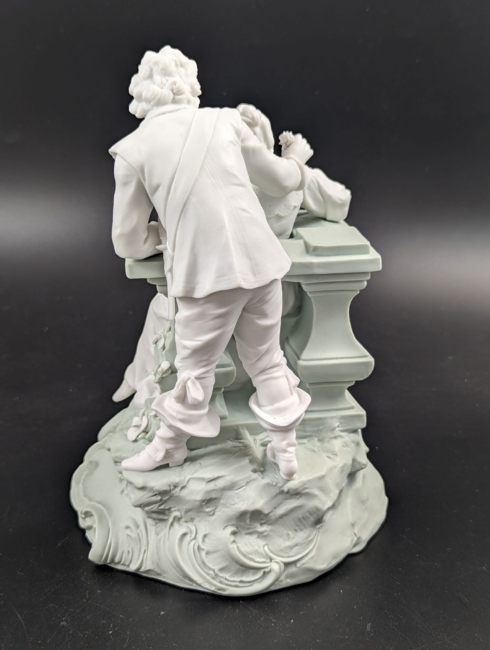 A white and pale green parian porcelain figurine depicting a courtly man and woman interacting beside a balustrade. View IV