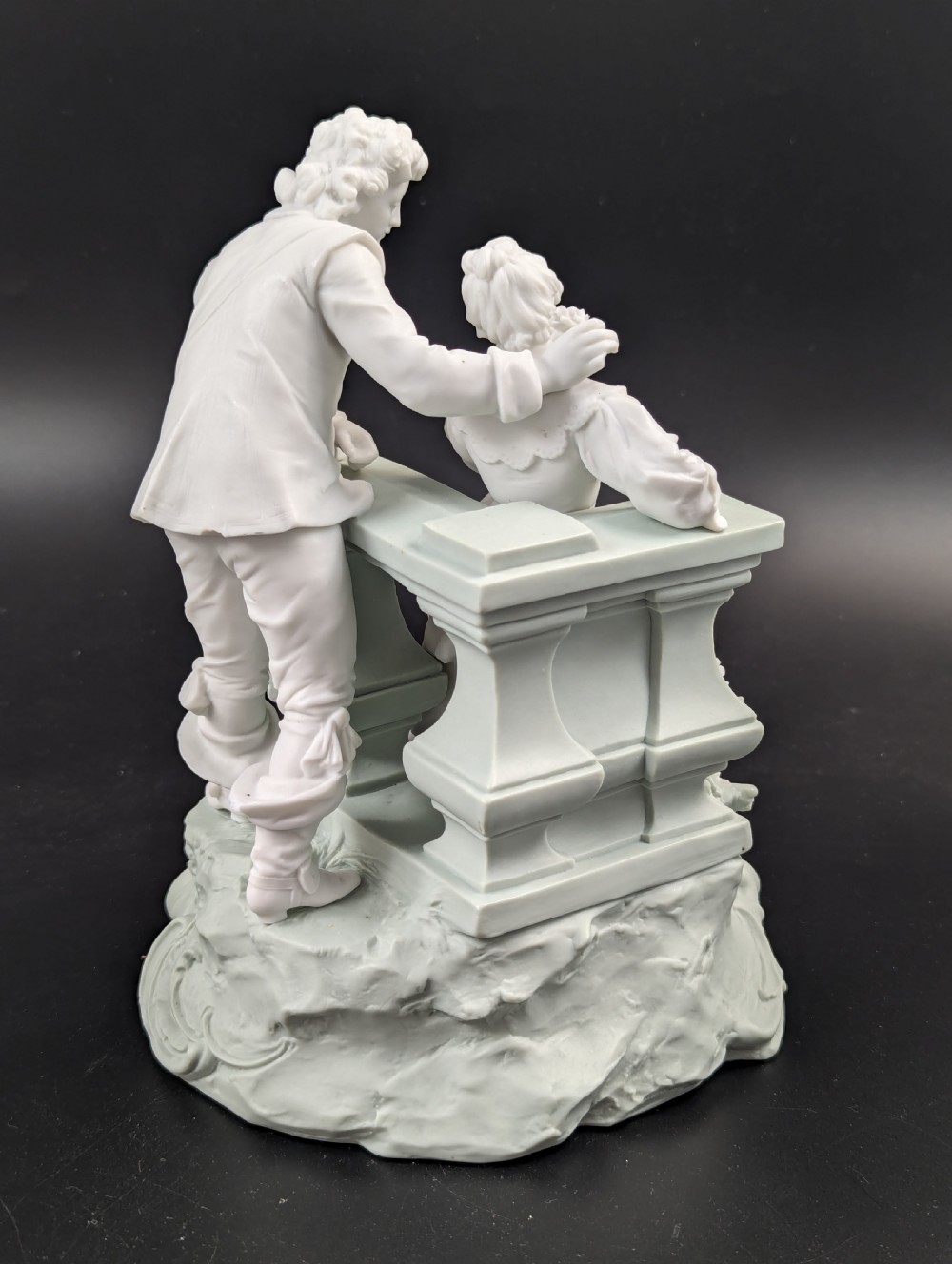 A white and pale green parian porcelain figurine depicting a courtly man and woman interacting beside a balustrade. View V