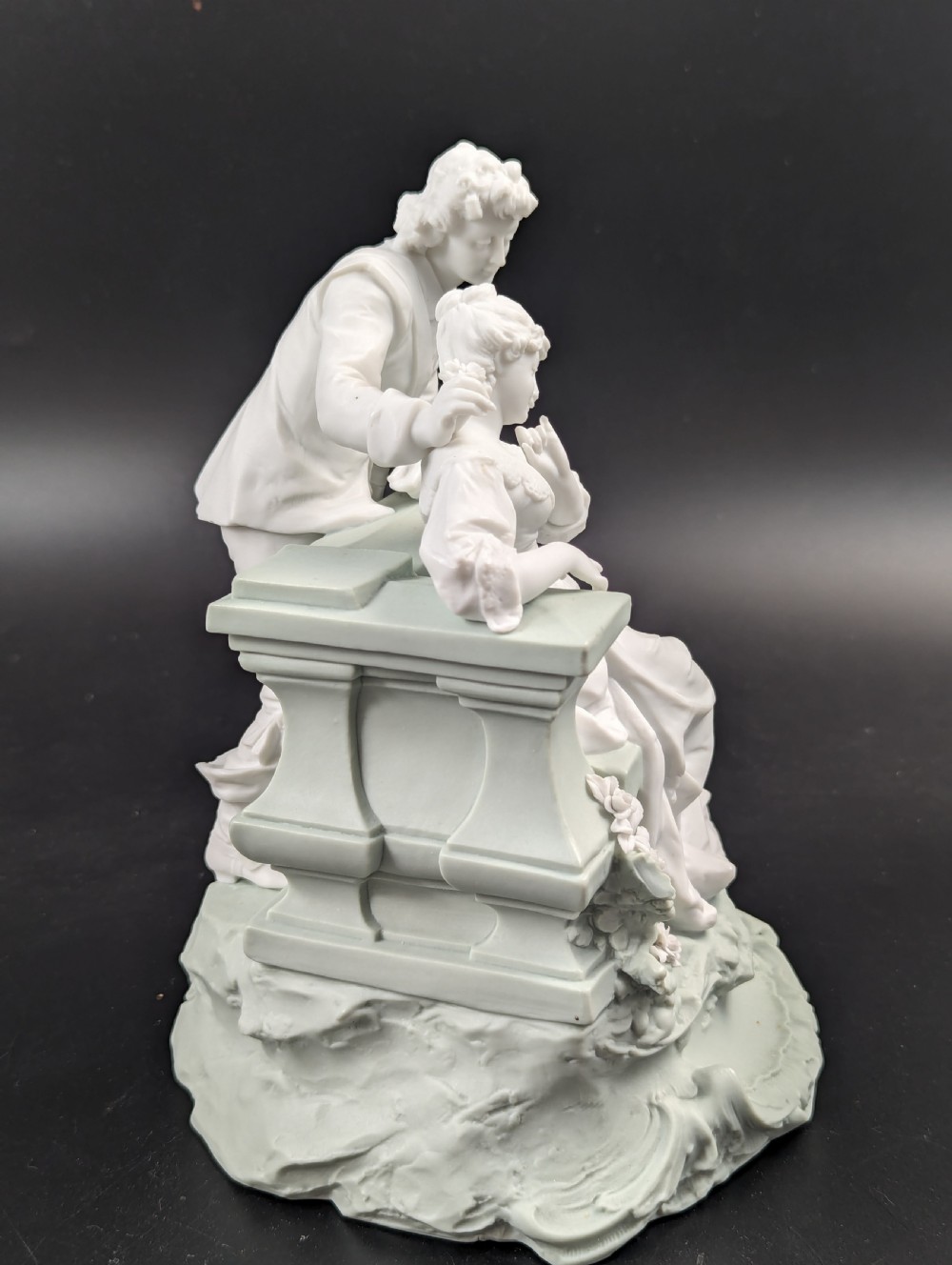 A white and pale green parian porcelain figurine depicting a courtly man and woman interacting beside a balustrade. View VI