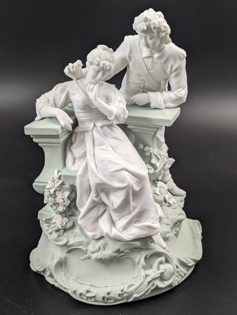 A white and pale green parian porcelain figurine depicting a courtly man and woman interacting beside a balustrade. View VIII