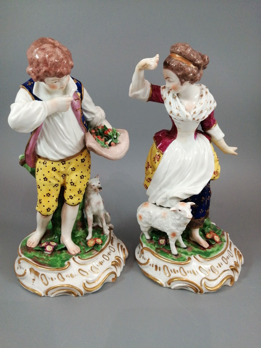 A pair of 19th-century French Samson porcelain figurines depicting a young boy with a basket of fruit and a dog alongside a shepherdess with a lamb, decorated with polychrome enamels and gilded scroll bases.