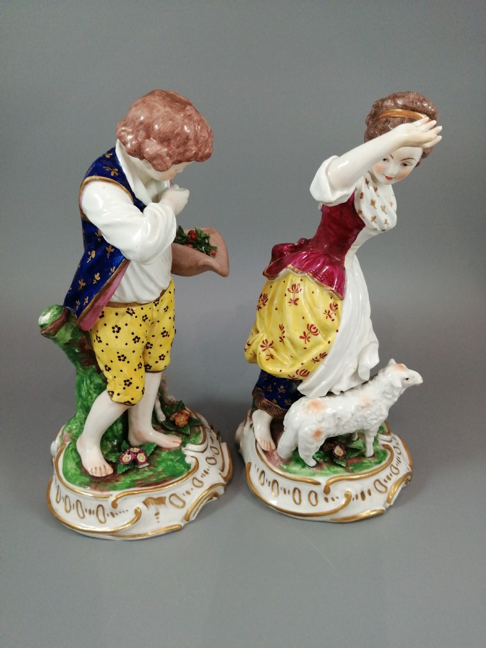 A pair of 19th-century French Samson porcelain figurines depicting a young boy with a basket of fruit and a dog alongside a shepherdess with a lamb, decorated with polychrome enamels and gilded scroll bases. View V