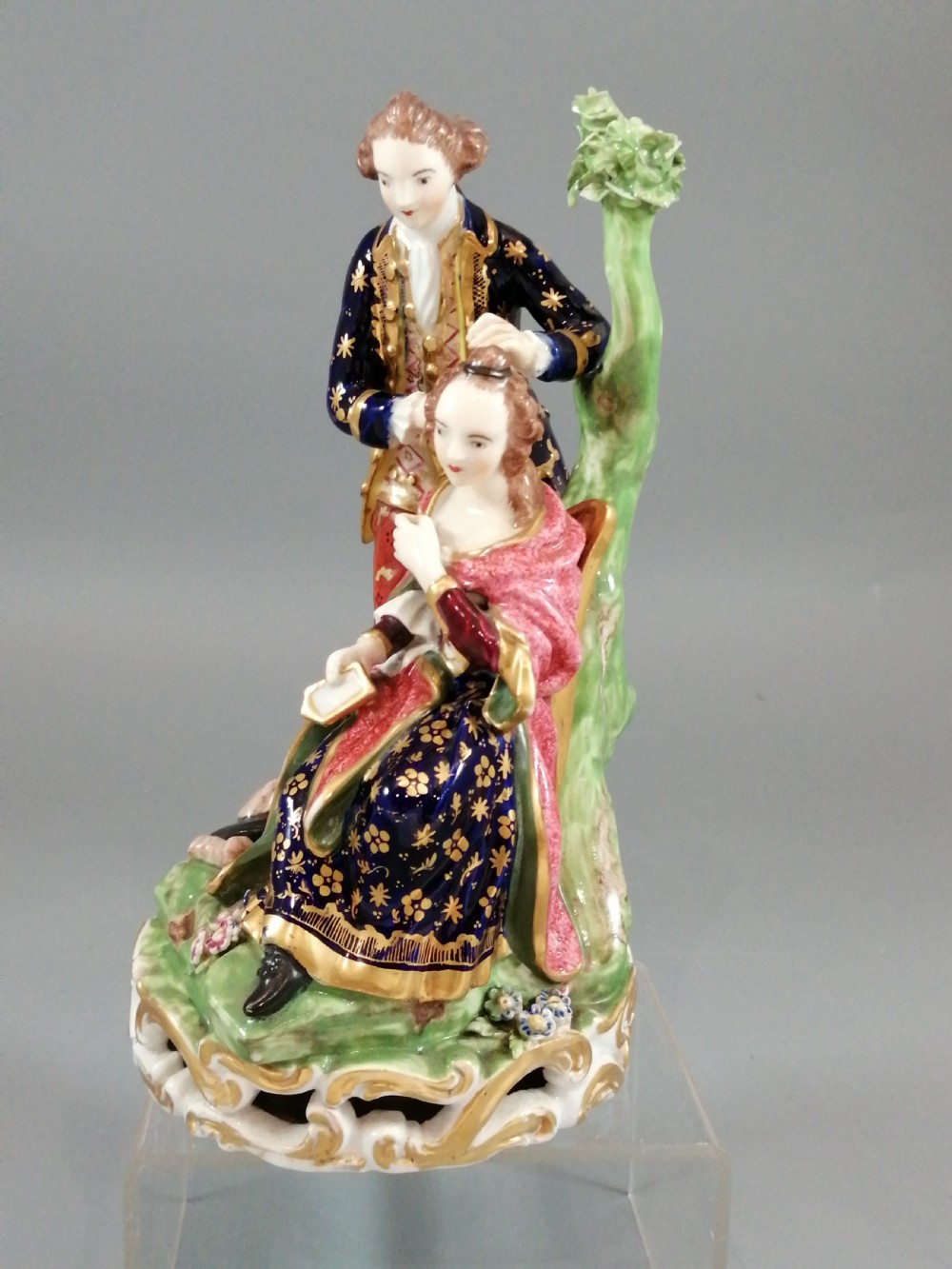 An early nineteenth-century painted porcelain figurine depicting a male court hairdresser styling a seated ladys hair on a rococo scroll base. View II