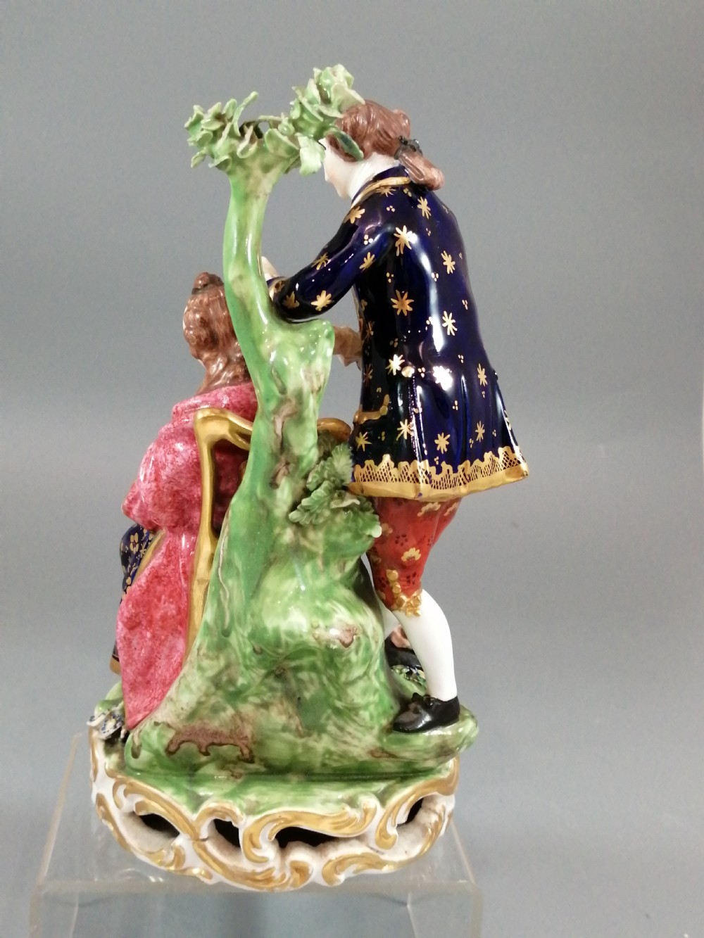 An early nineteenth-century painted porcelain figurine depicting a male court hairdresser styling a seated ladys hair on a rococo scroll base. View IV