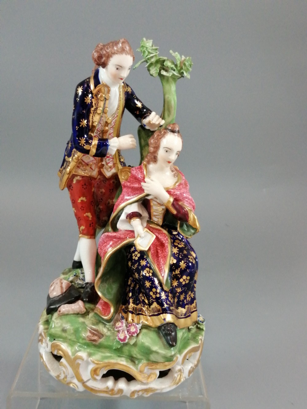 An early nineteenth-century painted porcelain figurine depicting a male court hairdresser styling a seated ladys hair on a rococo scroll base. View IX