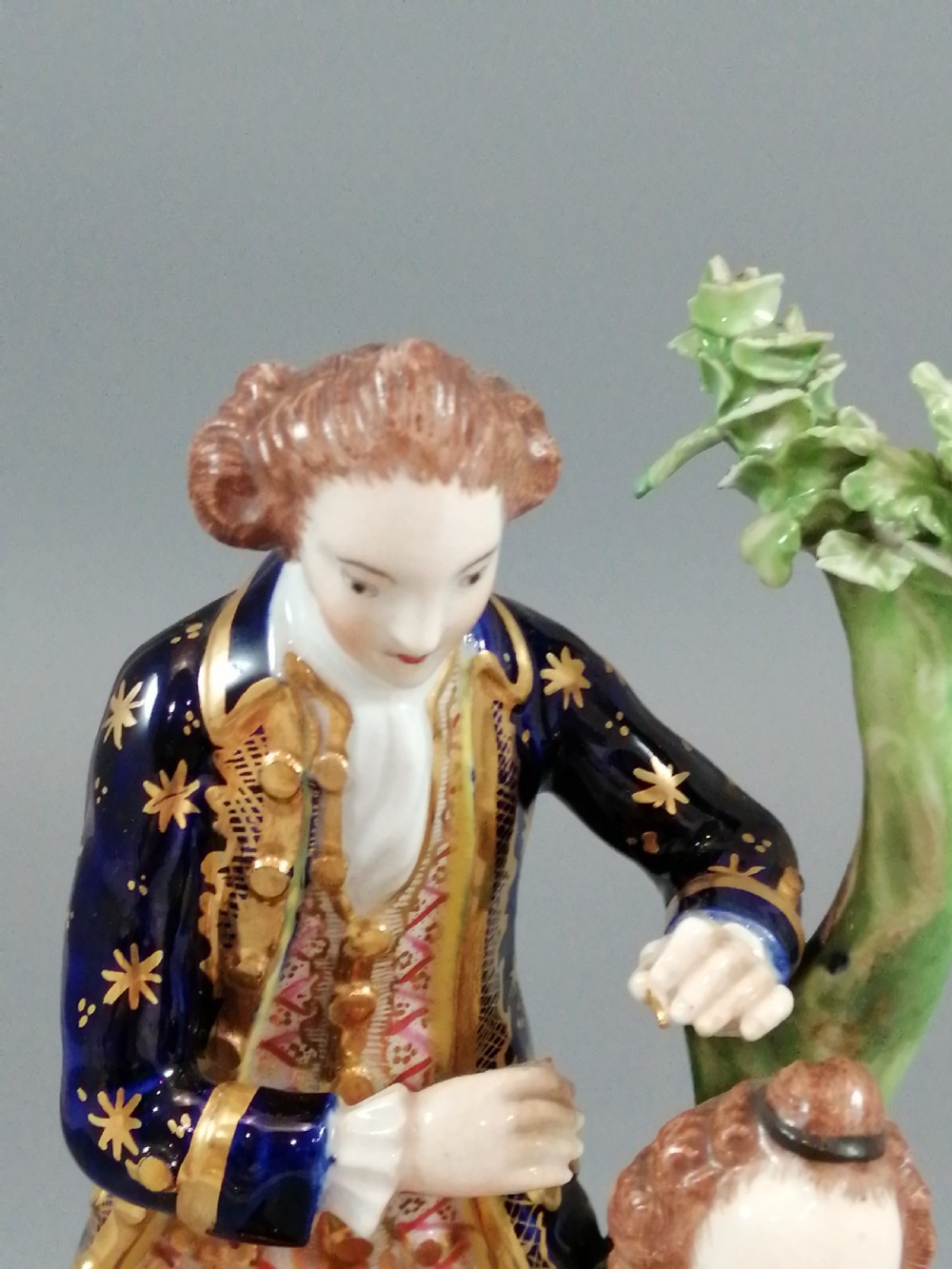An early nineteenth-century painted porcelain figurine depicting a male court hairdresser styling a seated ladys hair on a rococo scroll base. View XIV