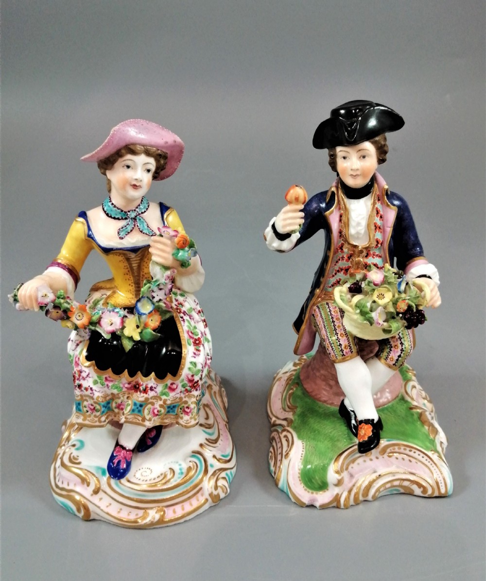Pair of hand-painted porcelain figurines depicting a male and female gardener seated on Rococo scrolling bases adorned with encrusted flowers.