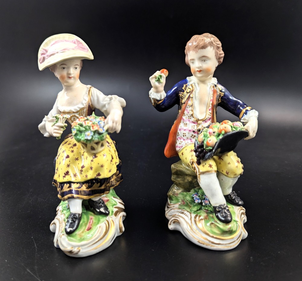 A pair of antique hand-painted Derby porcelain figurines featuring a young female gardener with a basket of flowers and a male gardener holding fruit on gilt scroll bases.