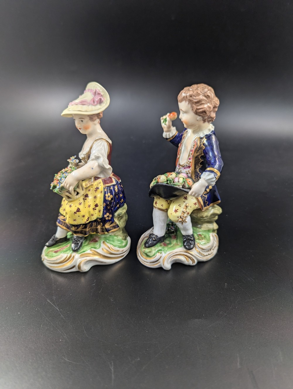 A pair of antique hand-painted Derby porcelain figurines featuring a young female gardener with a basket of flowers and a male gardener holding fruit on gilt scroll bases. View II