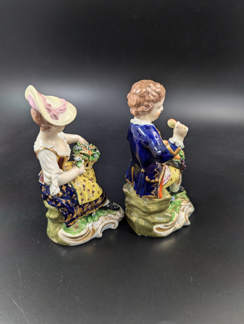 A pair of antique hand-painted Derby porcelain figurines featuring a young female gardener with a basket of flowers and a male gardener holding fruit on gilt scroll bases. View IV