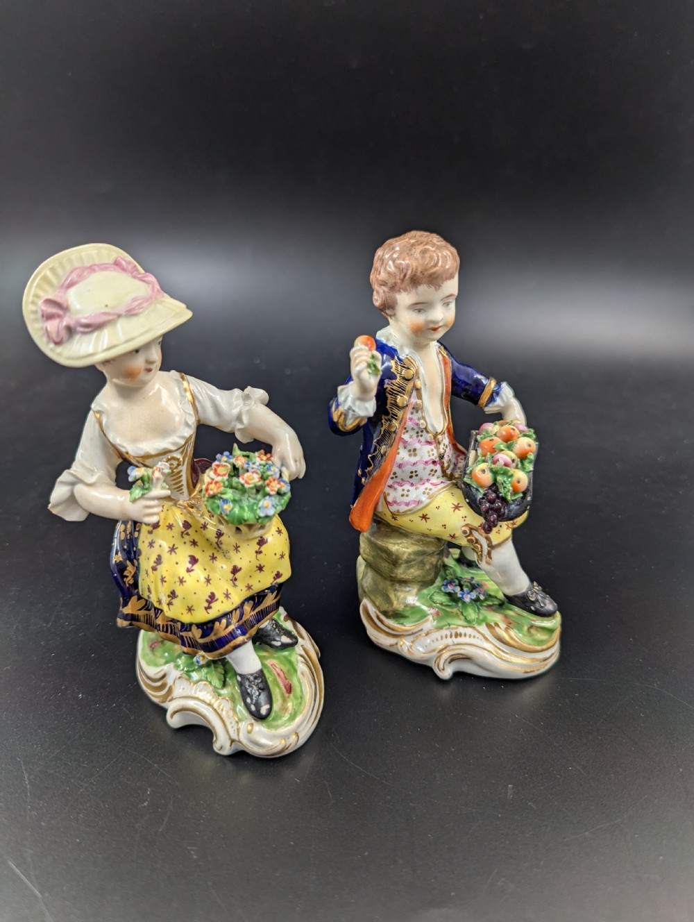 A pair of antique hand-painted Derby porcelain figurines featuring a young female gardener with a basket of flowers and a male gardener holding fruit on gilt scroll bases. View V