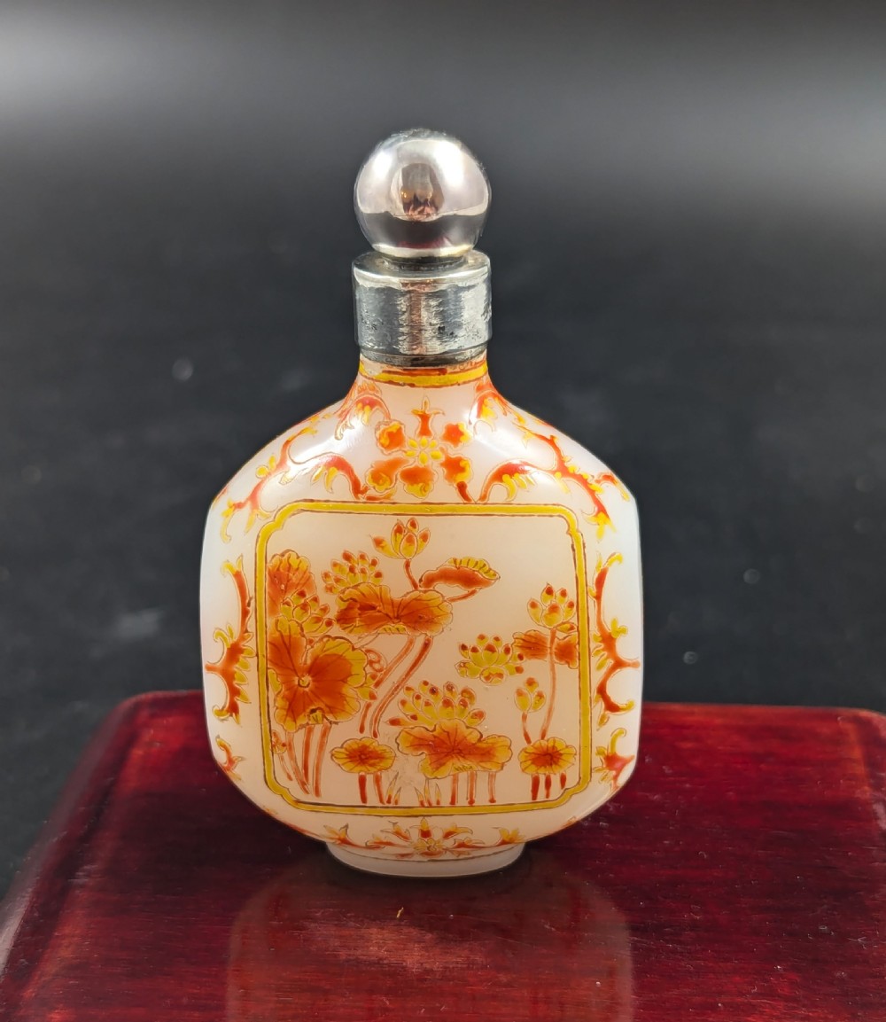 An opaque white glass snuff bottle featuring orange lotus enamel decoration within a central panel and a silver-tone spherical stopper.