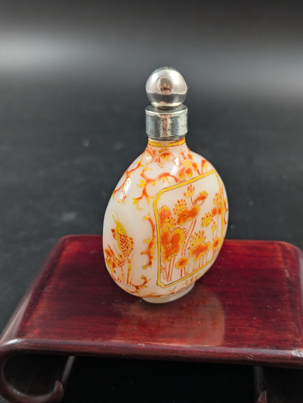 An opaque white glass snuff bottle featuring orange lotus enamel decoration within a central panel and a silver-tone spherical stopper. View VII