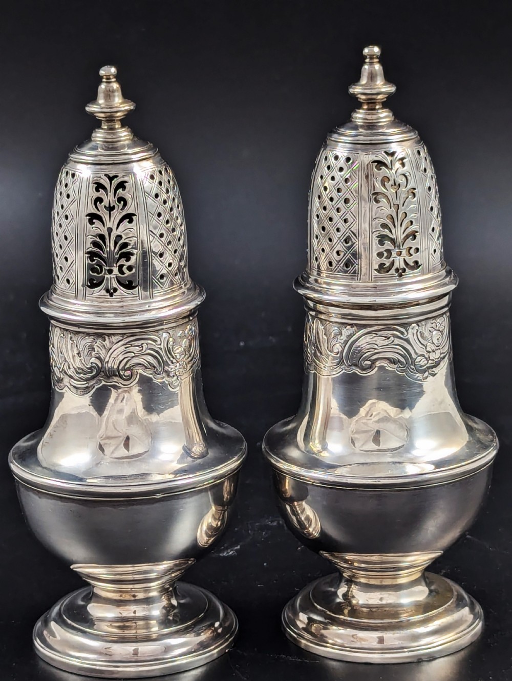 A pair of George II sterling silver baluster-form casters featuring pierced lattice tops, chased scroll detailing, and stepped pedestal bases.