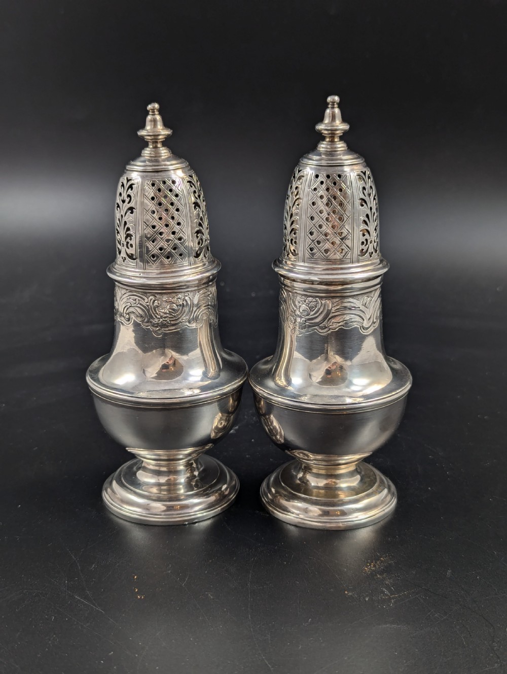 A pair of George II sterling silver baluster-form casters featuring pierced lattice tops, chased scroll detailing, and stepped pedestal bases. View III