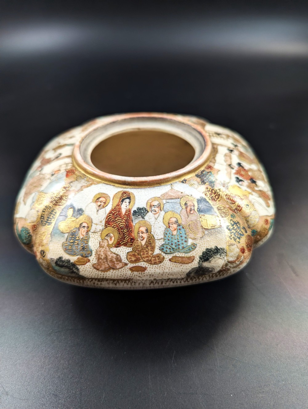 A squarish, lobed Satsuma earthenware vessel featuring hand-painted figures with gold halos, elaborate gilded borders, and a crackled ivory glaze.