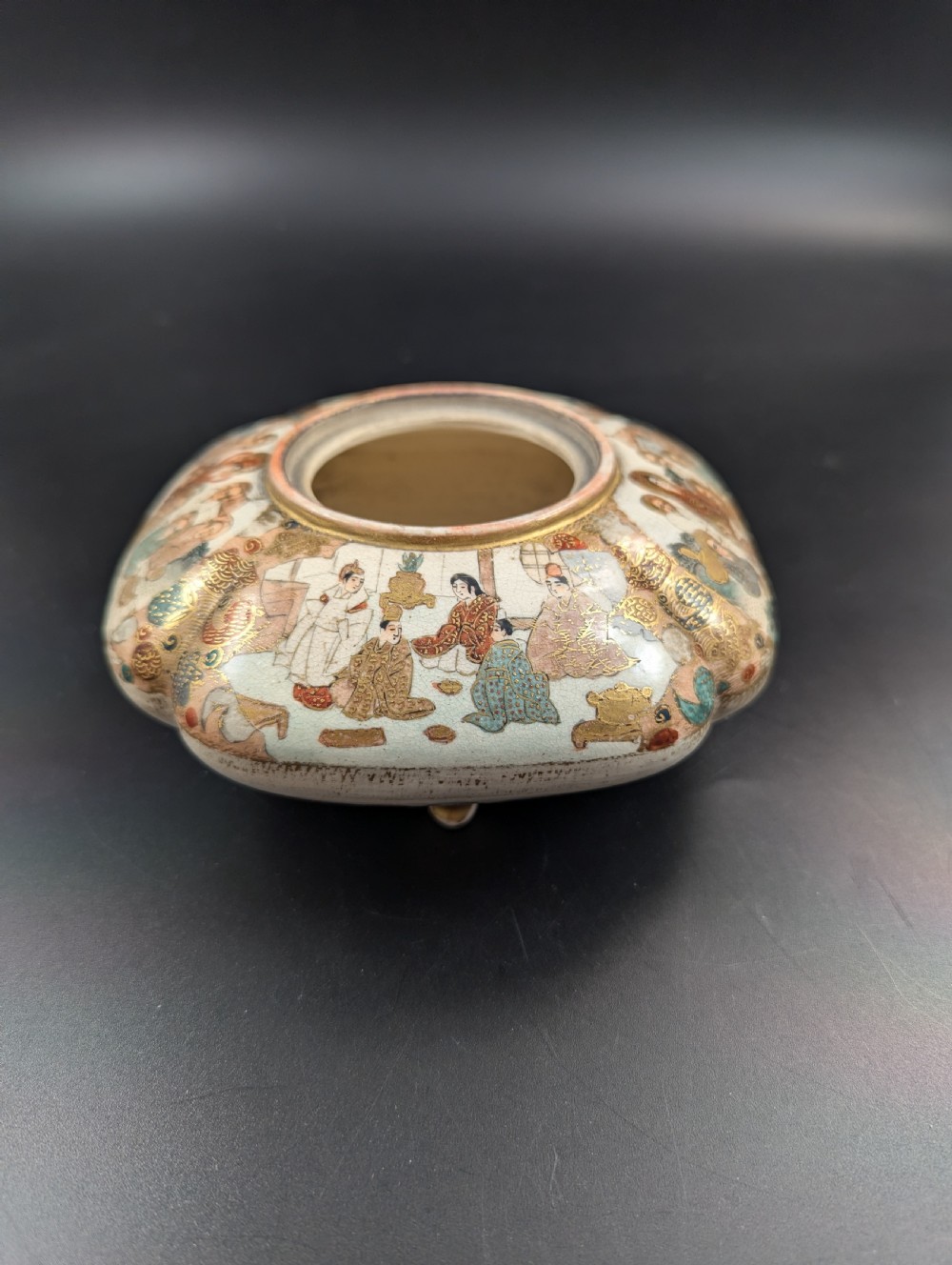 A squarish, lobed Satsuma earthenware vessel featuring hand-painted figures with gold halos, elaborate gilded borders, and a crackled ivory glaze. View VIII