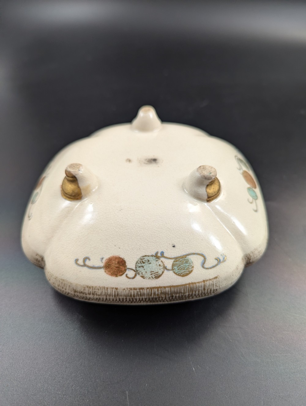 A squarish, lobed Satsuma earthenware vessel featuring hand-painted figures with gold halos, elaborate gilded borders, and a crackled ivory glaze. View XIV