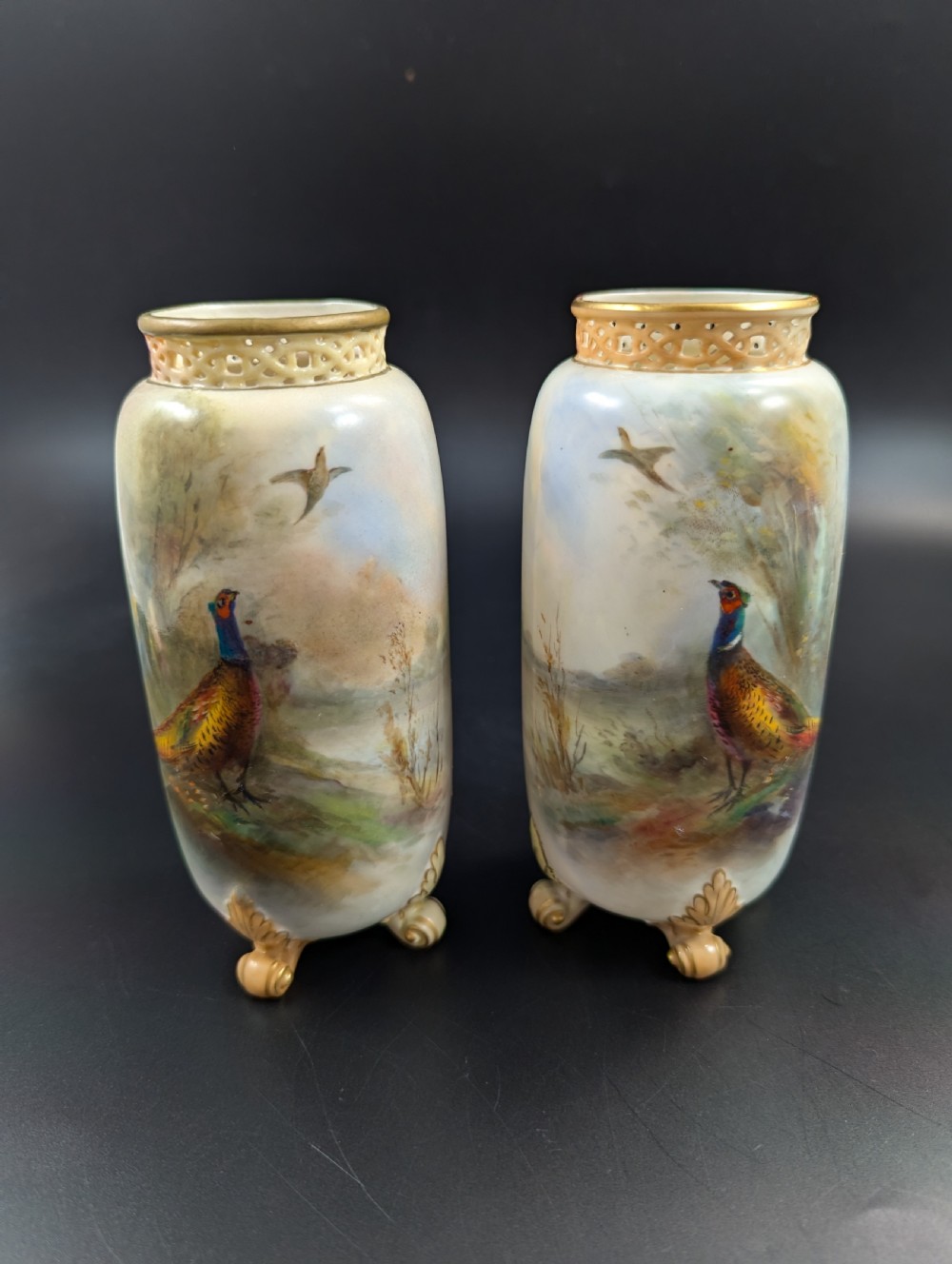 A pair of Edwardian Royal Worcester porcelain vases featuring hand-painted pheasants in natural landscapes, with reticulated rims and gilt scroll feet.