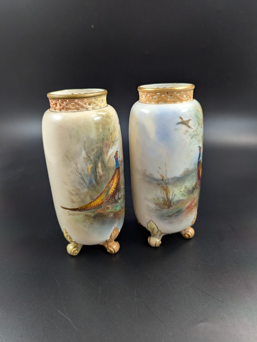 A pair of Edwardian Royal Worcester porcelain vases featuring hand-painted pheasants in natural landscapes, with reticulated rims and gilt scroll feet. View V