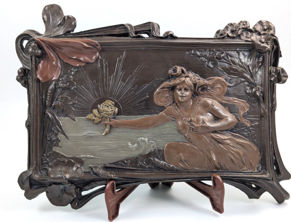 A rectangular French Art Nouveau terracotta relief featuring a draped female figure holding a rose against a rising sun background with a metallic finish.