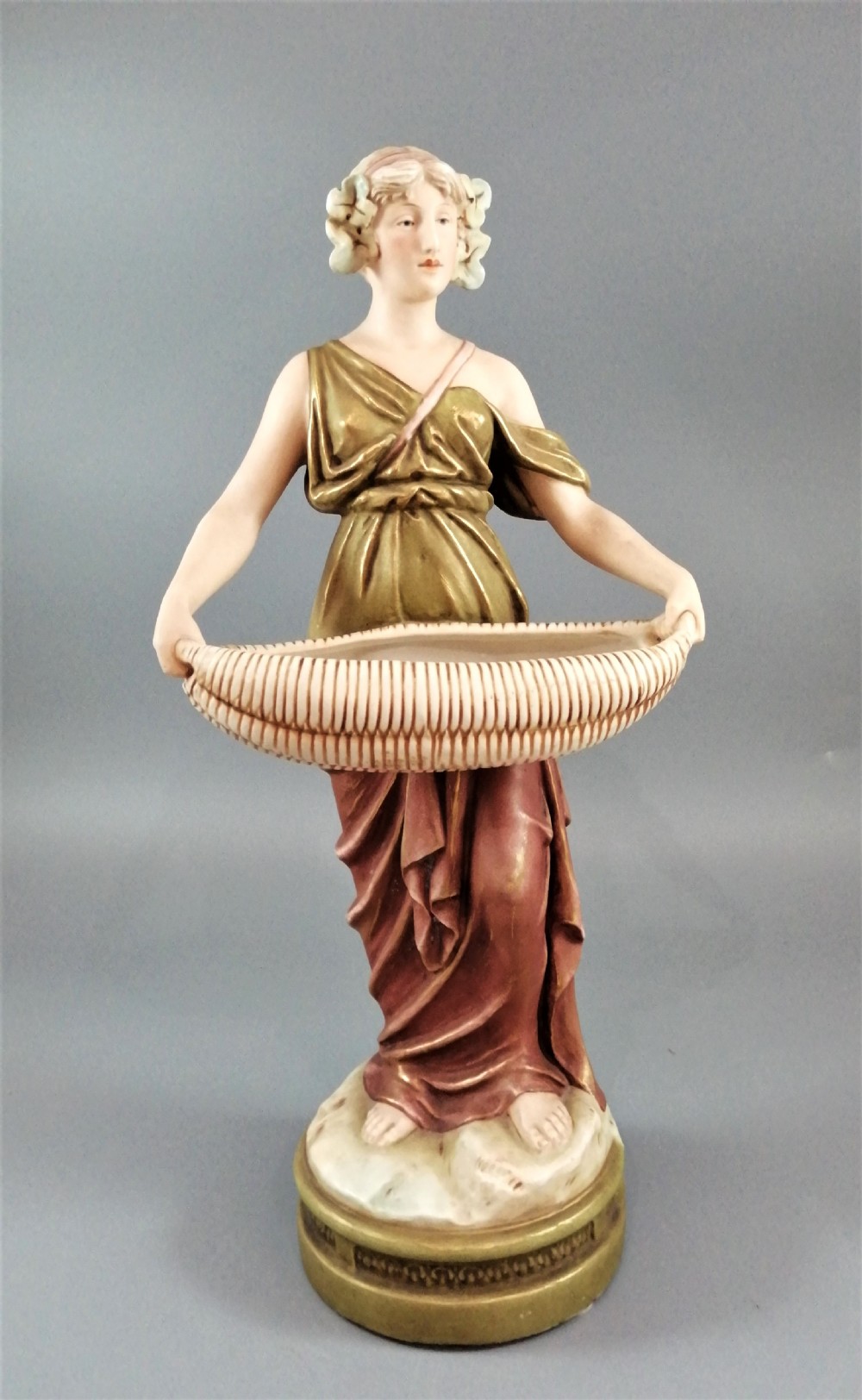 An Art Nouveau Royal Dux porcelain figurine depicting a draped maiden standing on a circular gilded pedestal holding a ribbed oval basket.