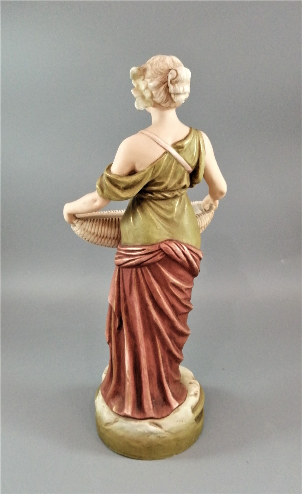 An Art Nouveau Royal Dux porcelain figurine depicting a draped maiden standing on a circular gilded pedestal holding a ribbed oval basket. View V
