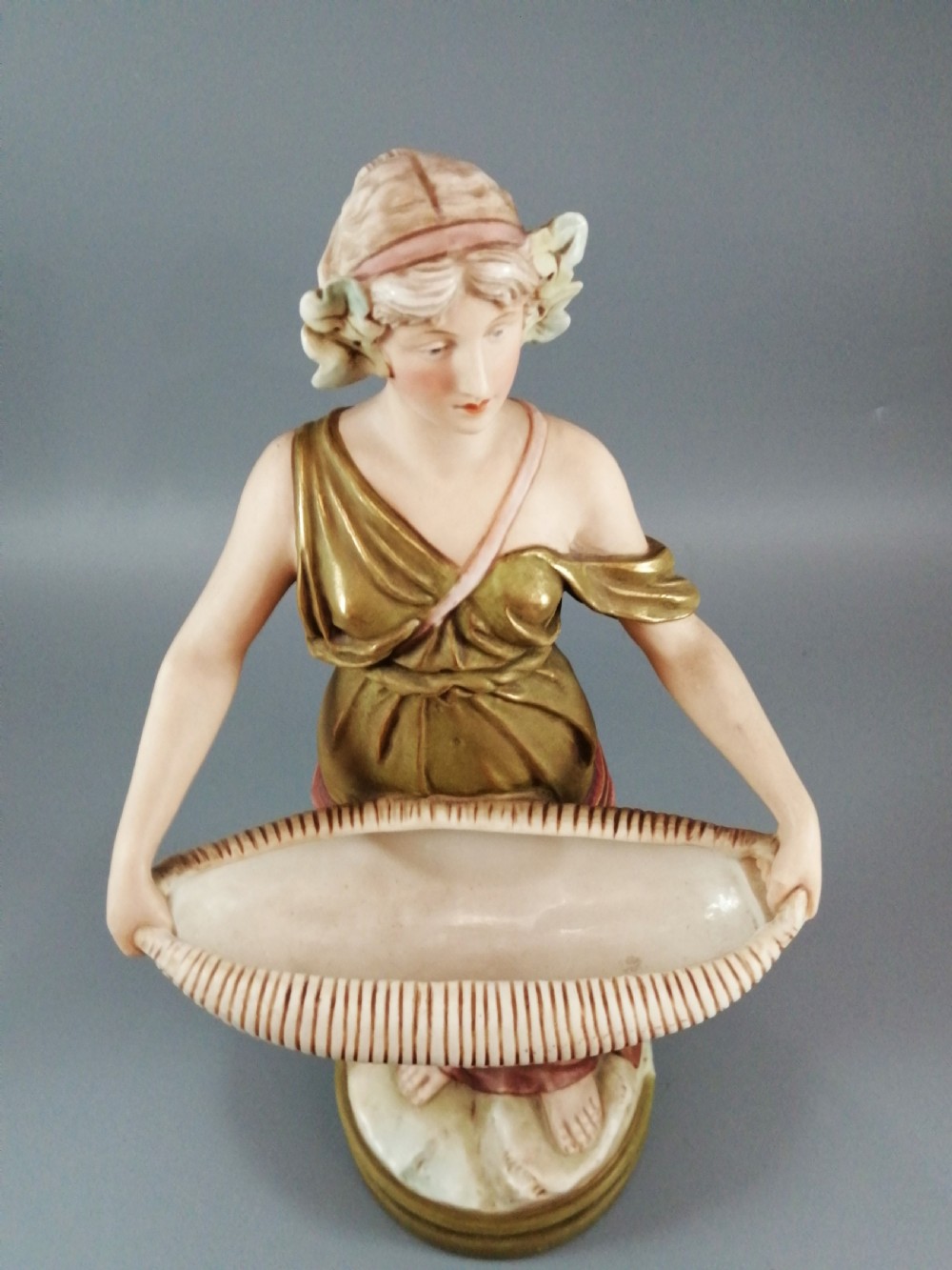 An Art Nouveau Royal Dux porcelain figurine depicting a draped maiden standing on a circular gilded pedestal holding a ribbed oval basket. View IX
