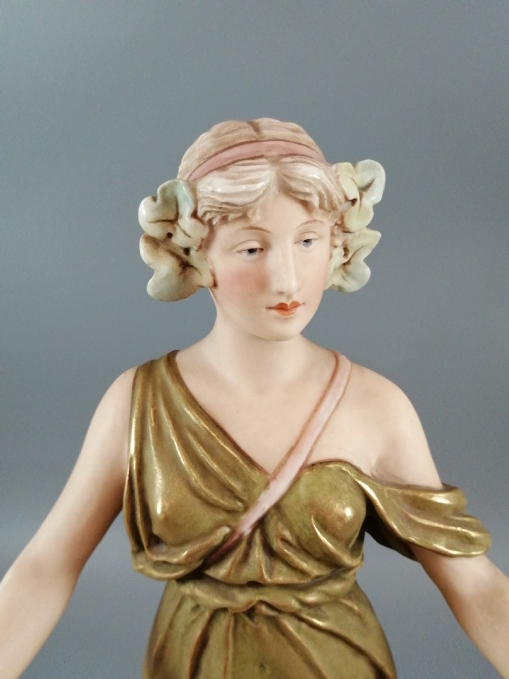 An Art Nouveau Royal Dux porcelain figurine depicting a draped maiden standing on a circular gilded pedestal holding a ribbed oval basket. View X
