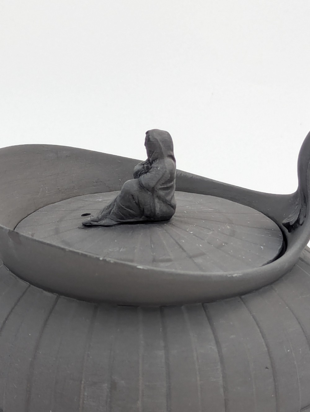 An eighteenth-century Wedgwood black basalt teapot featuring a ribbed stoneware body, high loop handle, and a seated female figure finial on the lid. View II