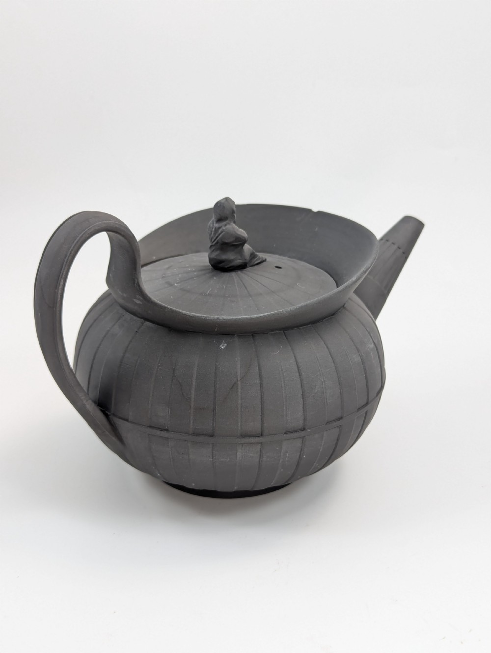 An eighteenth-century Wedgwood black basalt teapot featuring a ribbed stoneware body, high loop handle, and a seated female figure finial on the lid. View IV