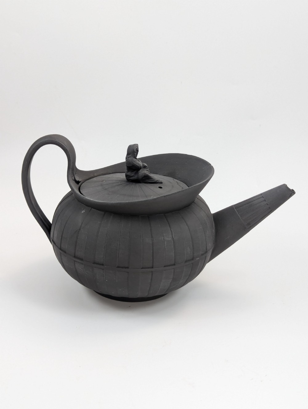 An eighteenth-century Wedgwood black basalt teapot featuring a ribbed stoneware body, high loop handle, and a seated female figure finial on the lid. View V