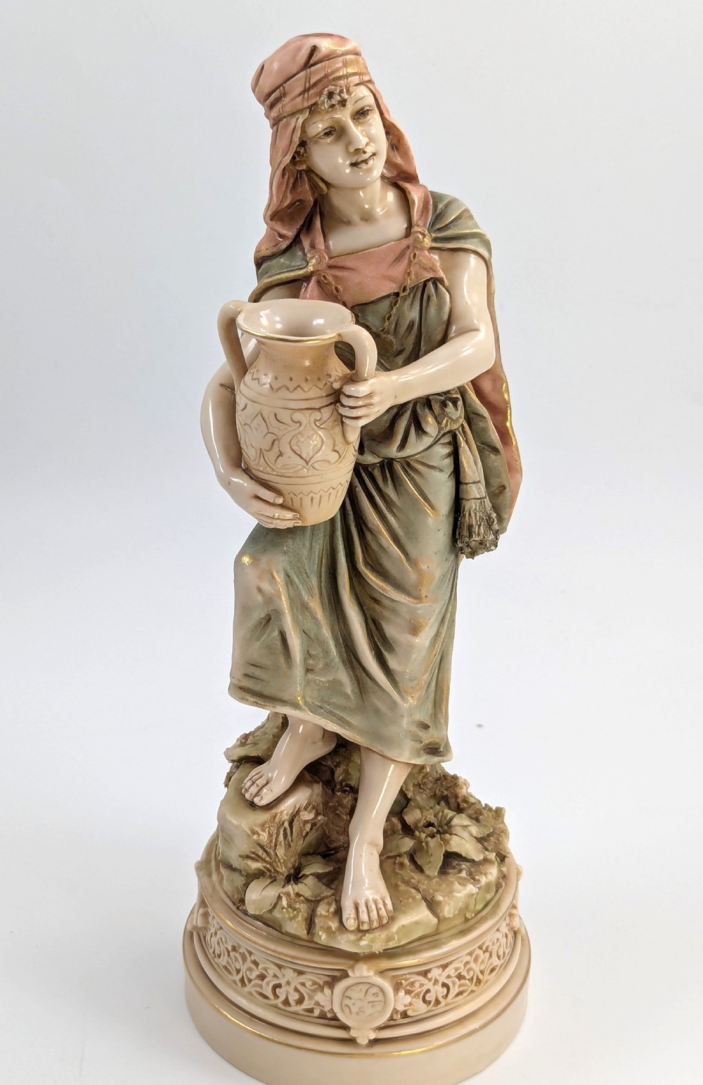 A porcelain figurine of a woman in draped robes carrying a double-handled water vessel on a patterned pedestal base.