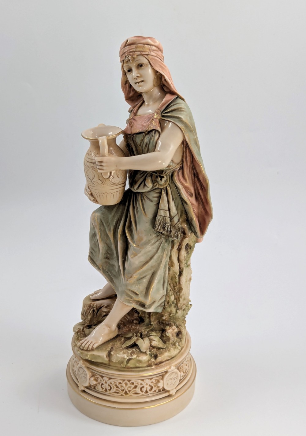 A porcelain figurine of a woman in draped robes carrying a double-handled water vessel on a patterned pedestal base. View II