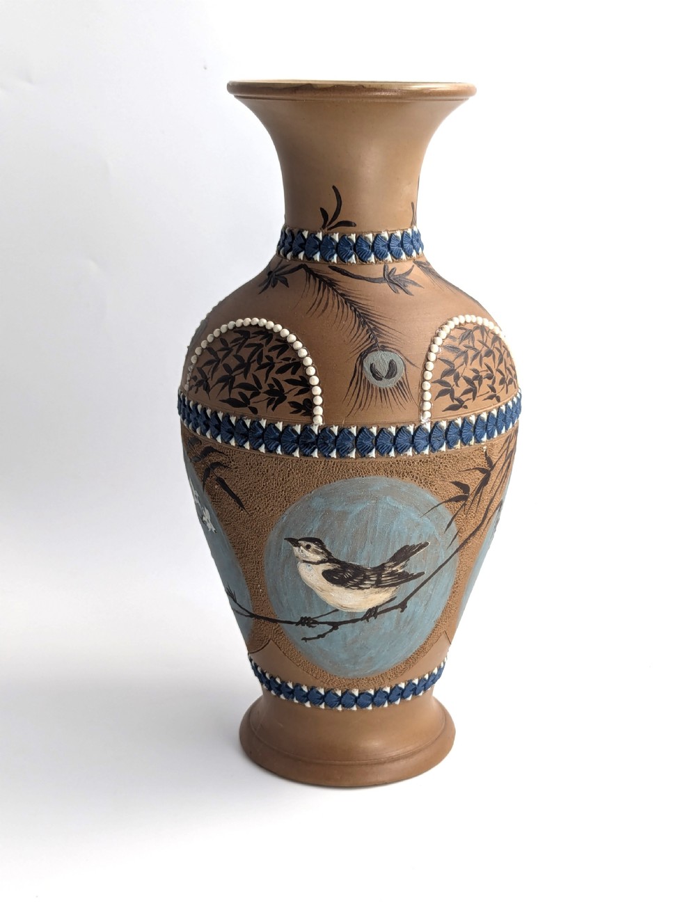A late 19th-century stoneware vase featuring a flared rim, warm brown buff body decorated with raised blue and white beaded borders, peacock feather motifs, and a painted central oval panel displaying a bird.