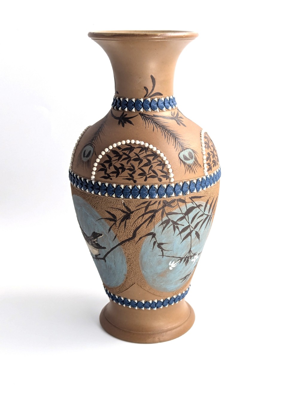 A late 19th-century stoneware vase featuring a flared rim, warm brown buff body decorated with raised blue and white beaded borders, peacock feather motifs, and a painted central oval panel displaying a bird. View II