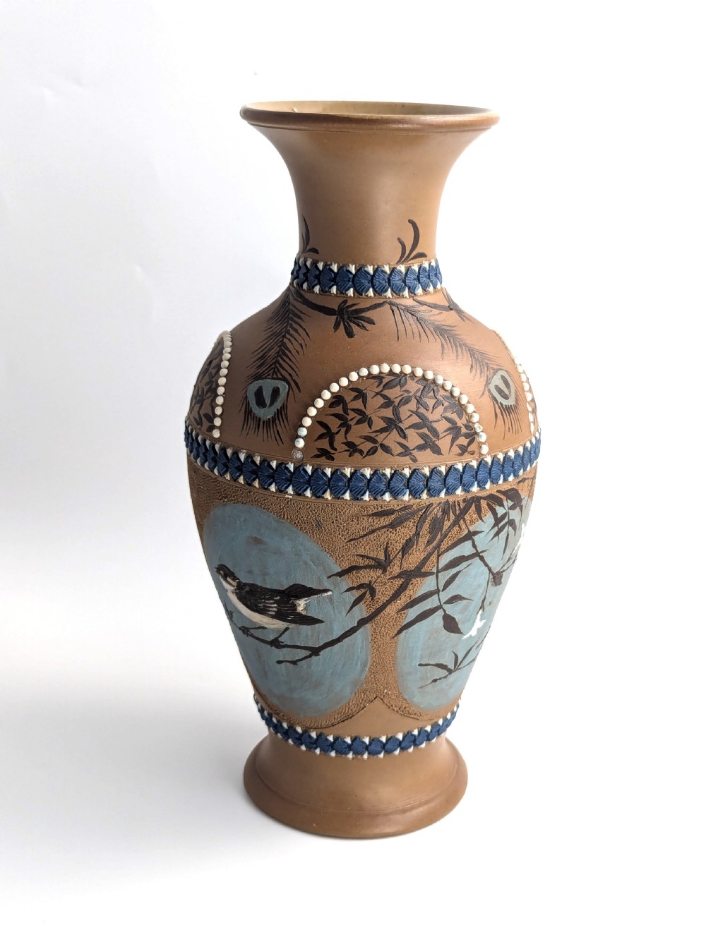 A late 19th-century stoneware vase featuring a flared rim, warm brown buff body decorated with raised blue and white beaded borders, peacock feather motifs, and a painted central oval panel displaying a bird. View IV