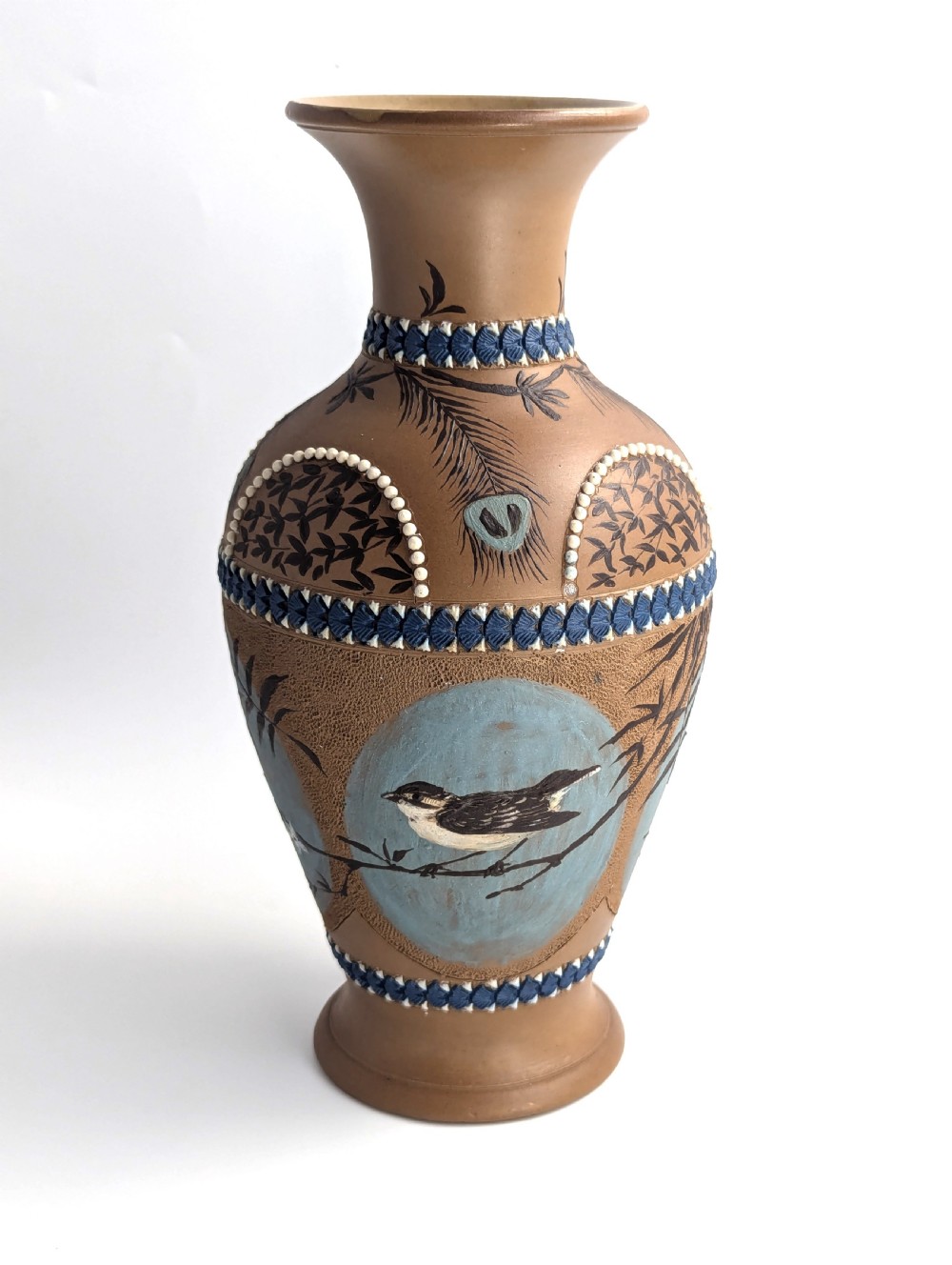 A late 19th-century stoneware vase featuring a flared rim, warm brown buff body decorated with raised blue and white beaded borders, peacock feather motifs, and a painted central oval panel displaying a bird. View V
