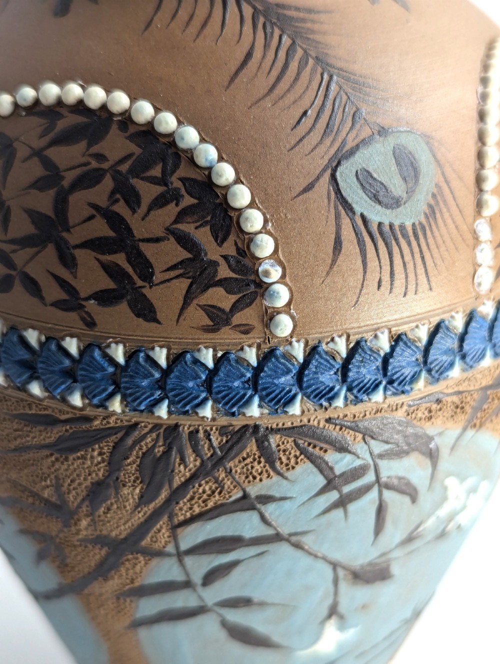 A late 19th-century stoneware vase featuring a flared rim, warm brown buff body decorated with raised blue and white beaded borders, peacock feather motifs, and a painted central oval panel displaying a bird. View XI