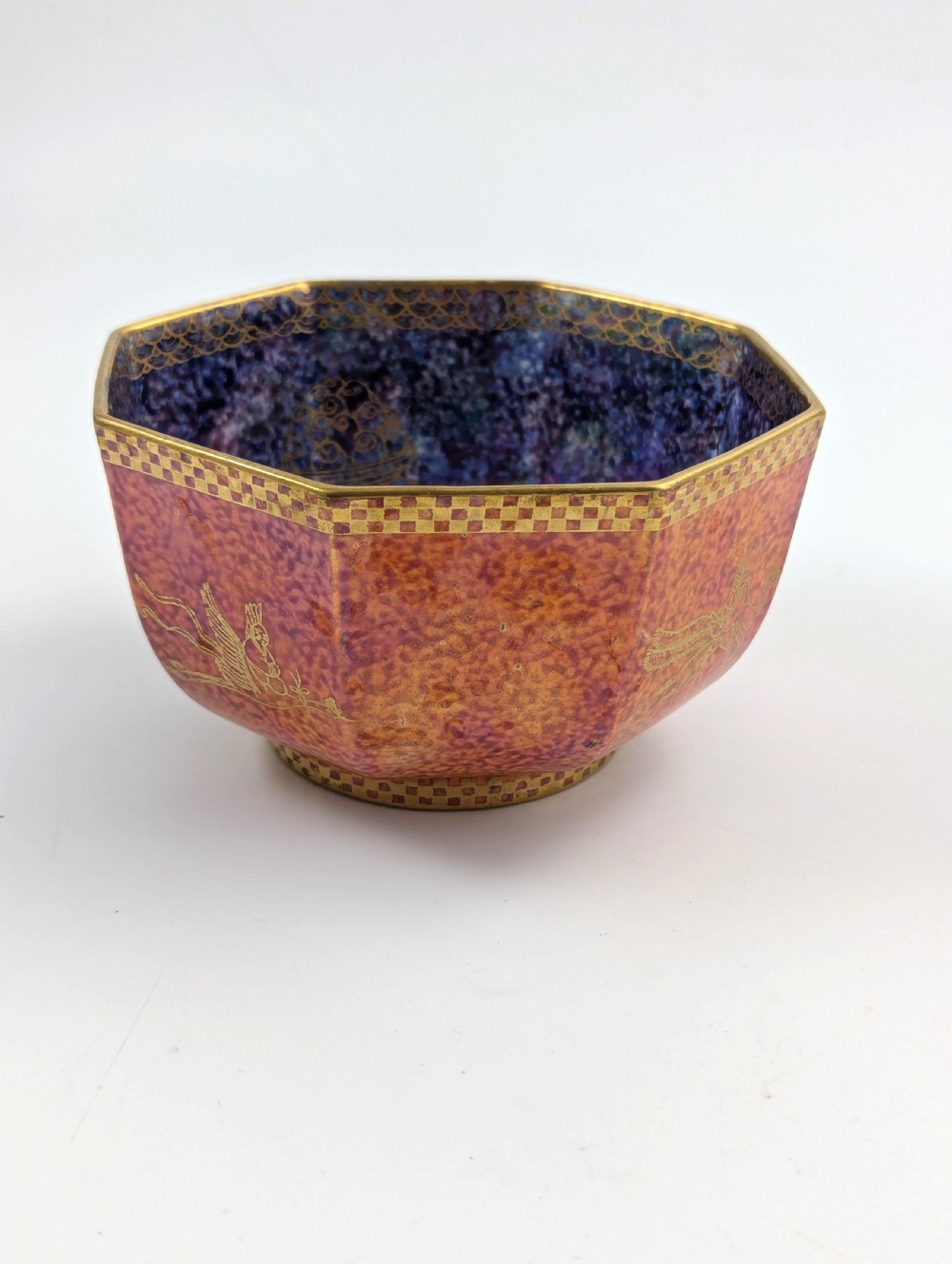 An octagonal Art Deco porcelain bowl featuring a mottled pink exterior with gold mythological motifs and an iridescent blue interior bounded by a gilded checkered rim. View II