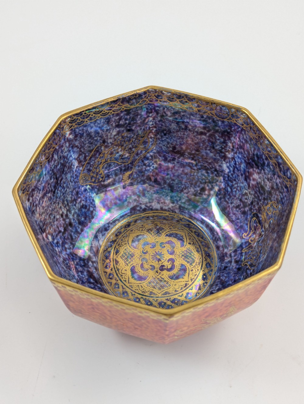 An octagonal Art Deco porcelain bowl featuring a mottled pink exterior with gold mythological motifs and an iridescent blue interior bounded by a gilded checkered rim. View VIII