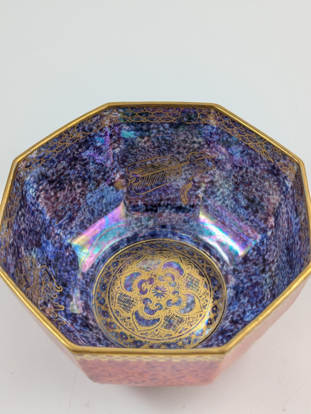 An octagonal Art Deco porcelain bowl featuring a mottled pink exterior with gold mythological motifs and an iridescent blue interior bounded by a gilded checkered rim. View XII