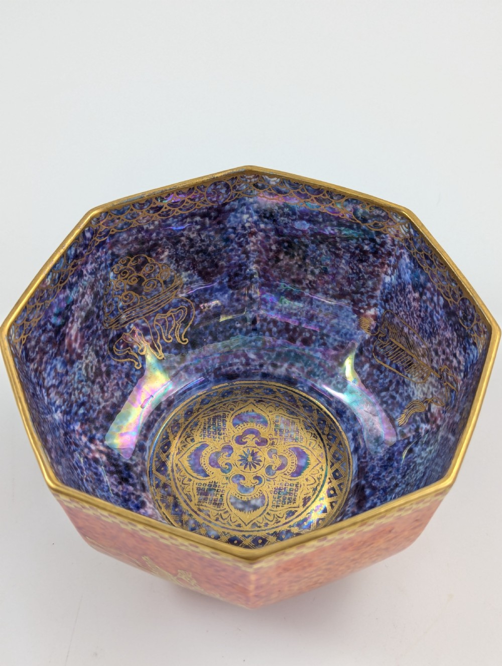 An octagonal Art Deco porcelain bowl featuring a mottled pink exterior with gold mythological motifs and an iridescent blue interior bounded by a gilded checkered rim. View XIII