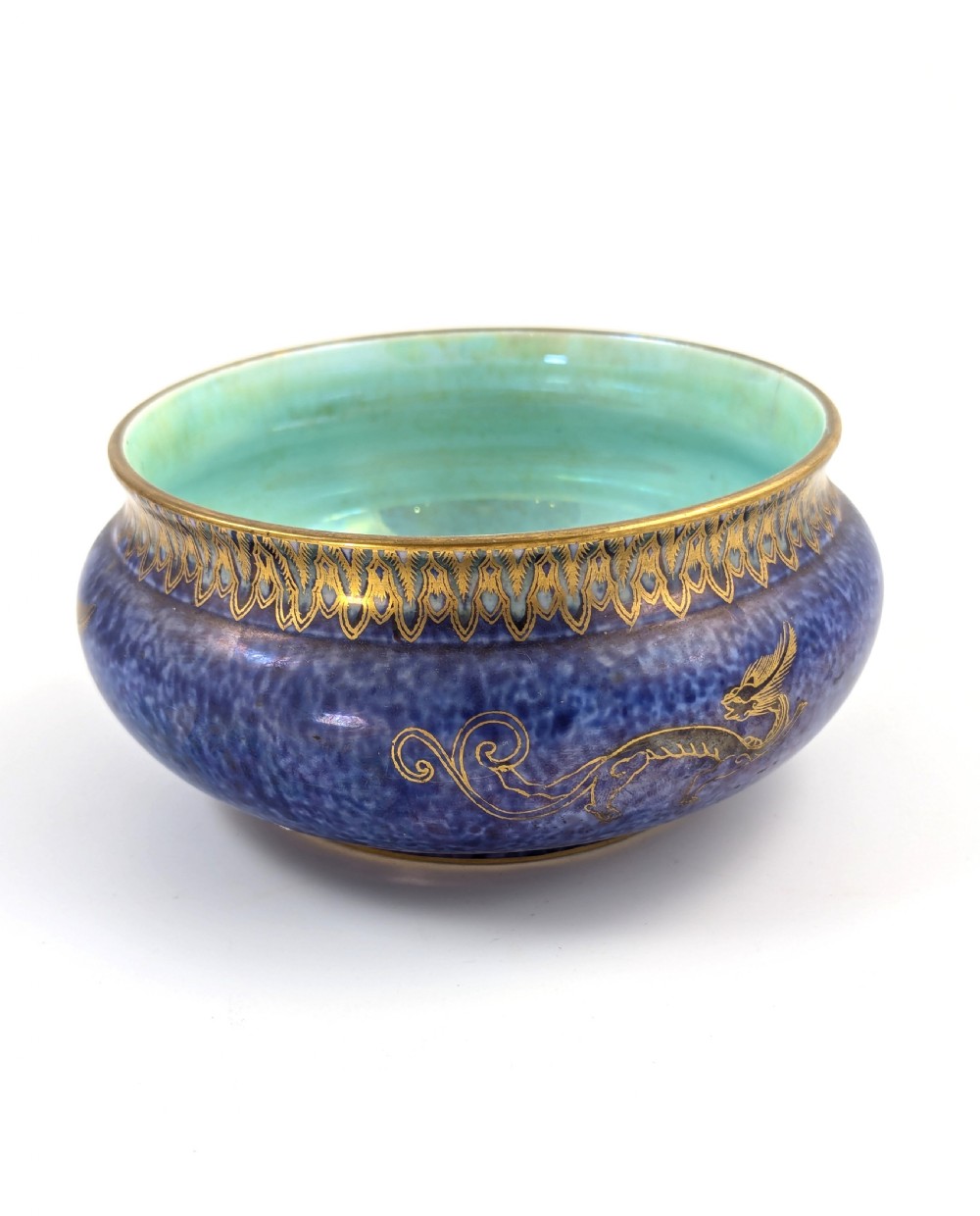 A rounded ceramic bowl featuring a mottled blue exterior decorated with a gilded dragon motif, a gilt patterned rim, and a pale iridescent green interior glaze.