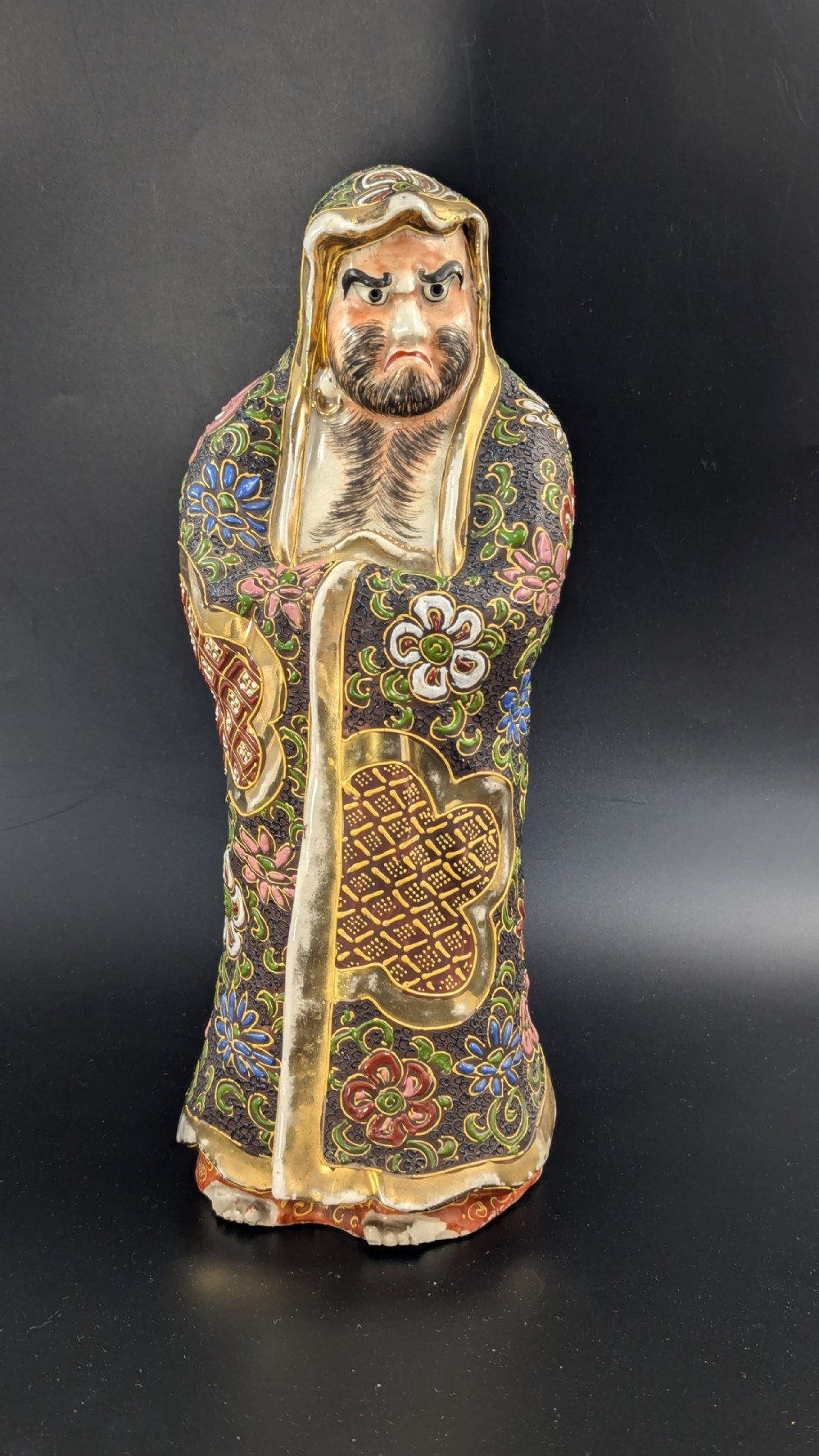 A Japanese Satsuma earthenware figurine depicting a bearded Daruma monk wearing a hooded robe with polychrome enamels and gold accents.