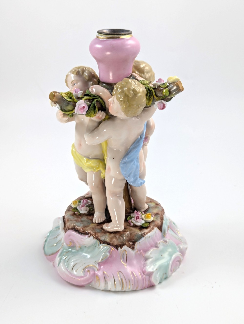 A 19th-century German hard-paste porcelain candlestick featuring two sculpted cherubs around a central column on a pastel Rococo scroll base. View II