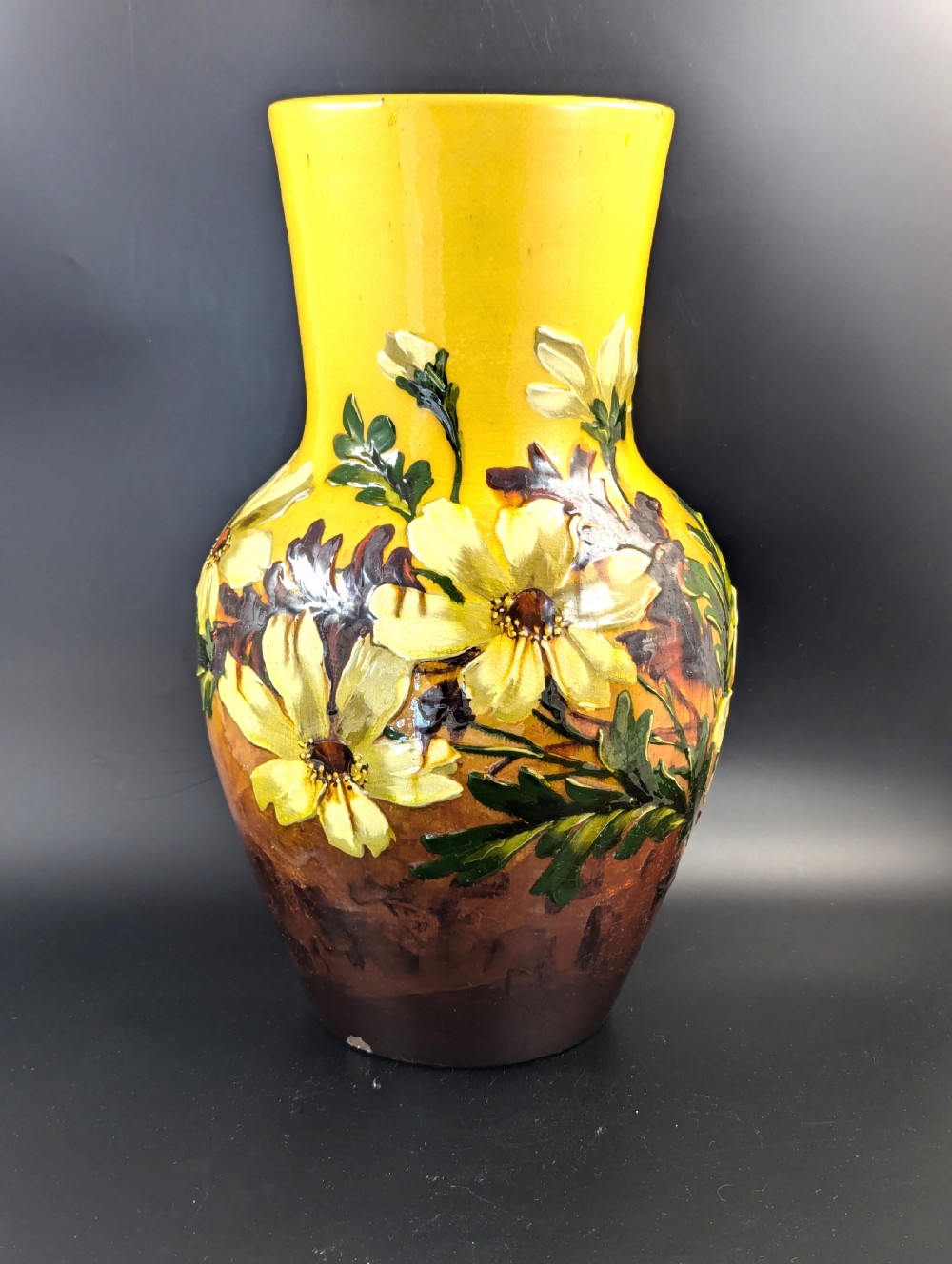 A tall ceramic vase featuring a yellow neck and upper body with raised impasto floral decoration transitioning into a dark brown glazed base.